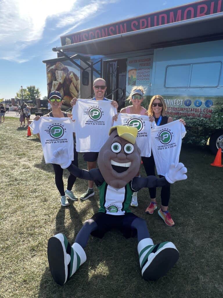 Four people stand smiling and holding Wisconsin Spudmobile T-shirts next to a large potato mascot in front of the Wisconsin Spudmobile truck at an outdoor event on a sunny day.